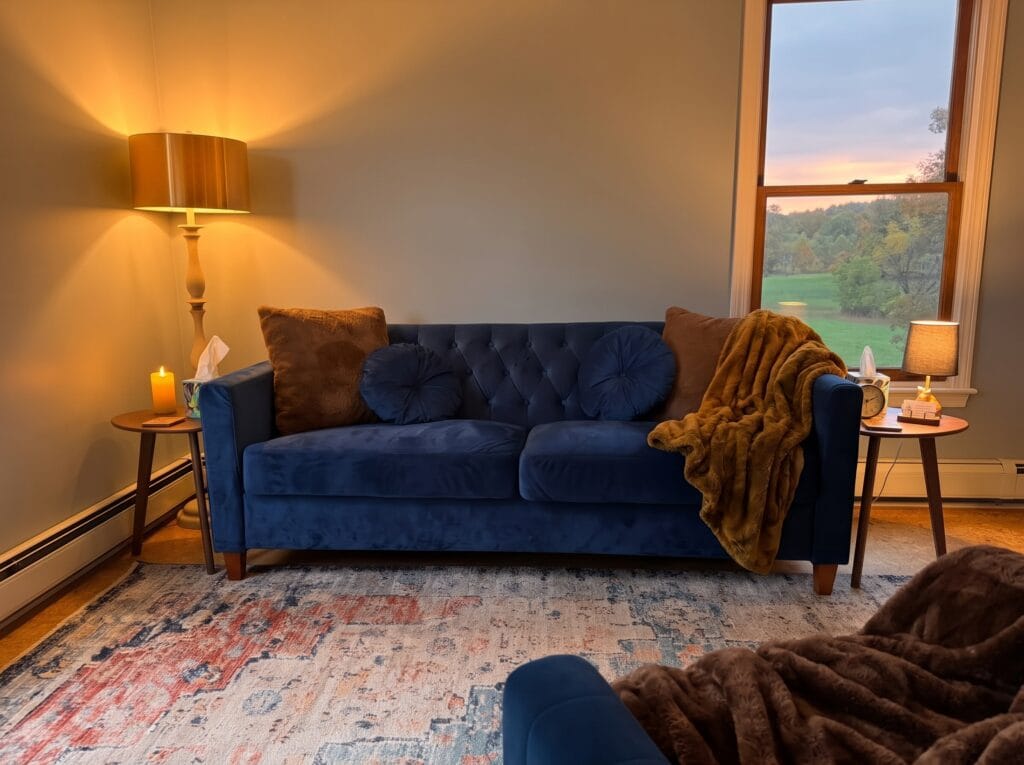 Navy velvet couch with a soft throw blanket, warm lamp light, and a window overlooking fields and trees at sunset, in Simon Abramson’s therapy office in Kingston, NY
